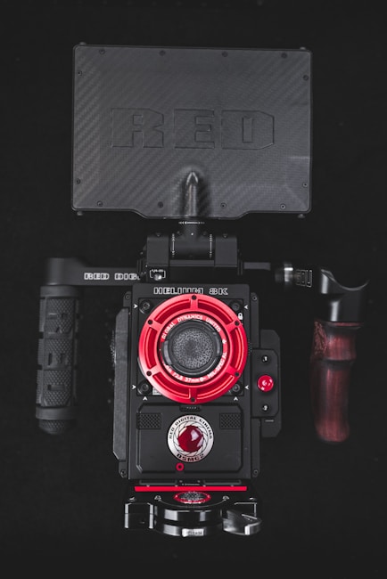 A professional camera with a prominent black and red design, featuring a camera body with multiple controls and a large screen on top. The front part includes a visible lens shielded by a detailed, industrial-looking red ring. The top part displays the brand name in large, embossed letters. Handles on either side appear sturdy, designed for secure grip, with one handle incorporating a wooden accent.