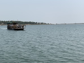 A ferry is carrying people and several motorcycles across a wide, calm river. The sky is clear and the distant shoreline is dotted with trees and cranes, suggesting a rural or semi-urban area.