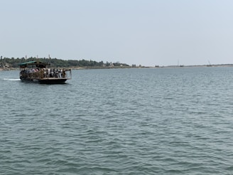 A ferry is carrying people and several motorcycles across a wide, calm river. The sky is clear and the distant shoreline is dotted with trees and cranes, suggesting a rural or semi-urban area.