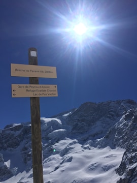 A wooden signpost with multiple directional signs stands in the foreground under a bright, sunlit sky. The signs point towards locations such as Brèche de Pacave, Gare de Peyrou d'Amont, Refuge Evariste Chancel, and Lac de Puy Vachier. Snow-covered mountains and a clear blue sky are visible in the background.