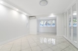 A spacious, empty room with white tiled flooring, walls, and ceiling. Large windows with shutters allow natural light to filter in. A mirrored wardrobe is positioned along one wall, and a ceiling light fixture provides additional illumination. An air conditioning unit is installed near the ceiling.