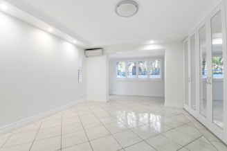 A spacious, empty room with white tiled flooring, walls, and ceiling. Large windows with shutters allow natural light to filter in. A mirrored wardrobe is positioned along one wall, and a ceiling light fixture provides additional illumination. An air conditioning unit is installed near the ceiling.