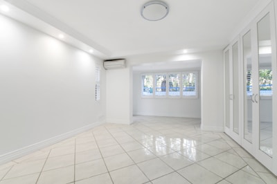 A spacious, empty room with white tiled flooring, walls, and ceiling. Large windows with shutters allow natural light to filter in. A mirrored wardrobe is positioned along one wall, and a ceiling light fixture provides additional illumination. An air conditioning unit is installed near the ceiling.