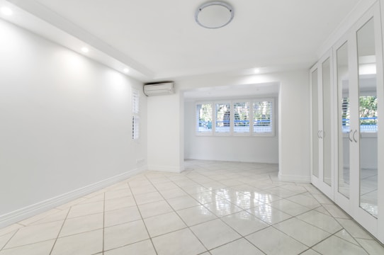 A spacious, empty room with white tiled flooring, walls, and ceiling. Large windows with shutters allow natural light to filter in. A mirrored wardrobe is positioned along one wall, and a ceiling light fixture provides additional illumination. An air conditioning unit is installed near the ceiling.