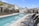 A residential area with modern townhouses surrounds a long, rectangular swimming pool. The pool has a clear blue hue, bordered by a textured concrete deck. Several dark wicker lounge chairs with armrests line one side of the pool, providing a place for relaxation. Young palm trees and ornamental plants are landscaped along the pool's edge. The sky is clear with a few small clouds, suggesting a sunny day.