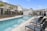 A residential area with modern townhouses surrounds a long, rectangular swimming pool. The pool has a clear blue hue, bordered by a textured concrete deck. Several dark wicker lounge chairs with armrests line one side of the pool, providing a place for relaxation. Young palm trees and ornamental plants are landscaped along the pool's edge. The sky is clear with a few small clouds, suggesting a sunny day.