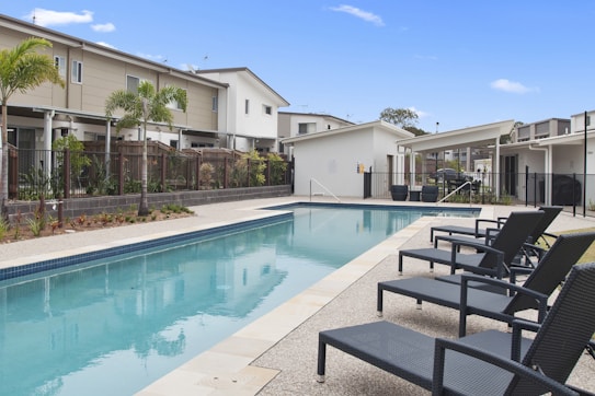 A residential area with modern townhouses surrounds a long, rectangular swimming pool. The pool has a clear blue hue, bordered by a textured concrete deck. Several dark wicker lounge chairs with armrests line one side of the pool, providing a place for relaxation. Young palm trees and ornamental plants are landscaped along the pool's edge. The sky is clear with a few small clouds, suggesting a sunny day.