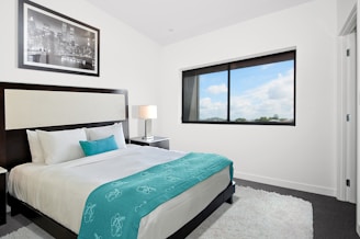 A contemporary bedroom featuring a large comfortable bed with white and teal bedding. A modern nightstand with a lamp is positioned beside the bed. A framed black and white cityscape photo adorns the wall above the headboard. A large window allows natural light to fill the room, offering a view of a partly cloudy sky. The room has a clean and minimalistic design, complemented by a soft, textured rug on the floor.