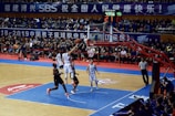 A basketball game takes place in a large indoor arena filled with spectators. Players from two teams, one in black uniforms and the other in white, compete for the ball near the hoop. The stadium is adorned with banners in Chinese, and a scoreboard displays a tied score of 21. The court features a distinctive blue and wood pattern.