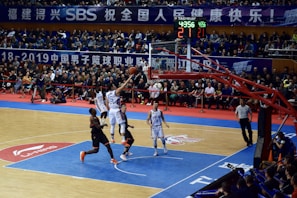 A basketball game takes place in a large indoor arena filled with spectators. Players from two teams, one in black uniforms and the other in white, compete for the ball near the hoop. The stadium is adorned with banners in Chinese, and a scoreboard displays a tied score of 21. The court features a distinctive blue and wood pattern.