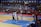 A basketball game takes place in a large indoor arena filled with spectators. Players from two teams, one in black uniforms and the other in white, compete for the ball near the hoop. The stadium is adorned with banners in Chinese, and a scoreboard displays a tied score of 21. The court features a distinctive blue and wood pattern.