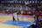 A basketball game takes place in a large indoor arena filled with spectators. Players from two teams, one in black uniforms and the other in white, compete for the ball near the hoop. The stadium is adorned with banners in Chinese, and a scoreboard displays a tied score of 21. The court features a distinctive blue and wood pattern.