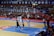 A basketball game takes place in a large indoor arena filled with spectators. Players from two teams, one in black uniforms and the other in white, compete for the ball near the hoop. The stadium is adorned with banners in Chinese, and a scoreboard displays a tied score of 21. The court features a distinctive blue and wood pattern.