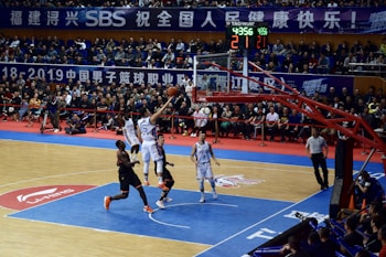 A basketball game takes place in a large indoor arena filled with spectators. Players from two teams, one in black uniforms and the other in white, compete for the ball near the hoop. The stadium is adorned with banners in Chinese, and a scoreboard displays a tied score of 21. The court features a distinctive blue and wood pattern.