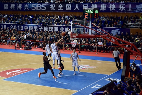 A basketball game takes place in a large indoor arena filled with spectators. Players from two teams, one in black uniforms and the other in white, compete for the ball near the hoop. The stadium is adorned with banners in Chinese, and a scoreboard displays a tied score of 21. The court features a distinctive blue and wood pattern.
