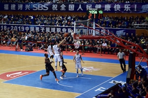 A basketball game takes place in a large indoor arena filled with spectators. Players from two teams, one in black uniforms and the other in white, compete for the ball near the hoop. The stadium is adorned with banners in Chinese, and a scoreboard displays a tied score of 21. The court features a distinctive blue and wood pattern.
