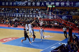 A basketball game takes place in a large indoor arena filled with spectators. Players from two teams, one in black uniforms and the other in white, compete for the ball near the hoop. The stadium is adorned with banners in Chinese, and a scoreboard displays a tied score of 21. The court features a distinctive blue and wood pattern.