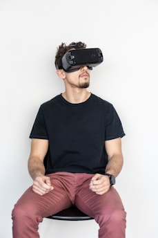 A person wearing a black virtual reality headset sits with a focused expression. They are dressed in a black t-shirt and reddish-brown pants and appear to be engaging with the virtual environment. Their hands are positioned as if holding invisible controls.