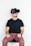 A person wearing a black virtual reality headset sits with a focused expression. They are dressed in a black t-shirt and reddish-brown pants and appear to be engaging with the virtual environment. Their hands are positioned as if holding invisible controls.
