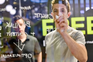 A person is writing on a transparent glass board with various words and diagrams visible. Another individual, slightly out of focus, is observing from the other side of the glass.