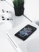 A smartphone with a colorful and futuristic display lies on top of a white gaming console. Two white gaming controllers are placed nearby on a white surface. In the background, there is a small black pot containing green, leafy plants, adding contrast and a touch of nature to the modern setup.