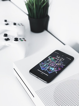 A smartphone with a colorful and futuristic display lies on top of a white gaming console. Two white gaming controllers are placed nearby on a white surface. In the background, there is a small black pot containing green, leafy plants, adding contrast and a touch of nature to the modern setup.