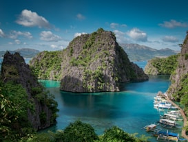 A serene tropical landscape featuring towering limestone cliffs covered in lush greenery surrounding a turquoise lagoon. Several traditional boats are docked at a wooden pier on the right side of the image. The sky is clear with a few scattered clouds, creating a picturesque and peaceful atmosphere.