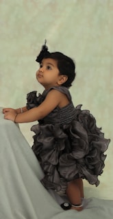 A young toddler wearing a frilly, dark-colored dress is gazing upward with an expression of curiosity. The child is standing against a soft, light-colored, textured background and appears to be holding onto an object draped in gray fabric.