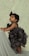 A young toddler wearing a frilly, dark-colored dress is gazing upward with an expression of curiosity. The child is standing against a soft, light-colored, textured background and appears to be holding onto an object draped in gray fabric.