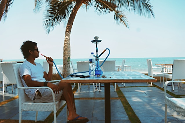 A person is sitting at a seaside cafe, enjoying a hookah. The setting includes palm trees, the ocean, and a blue sky. The individual, wearing a white shirt and shorts, relaxes in a wicker chair. A water bottle and a smartphone are placed on the table.