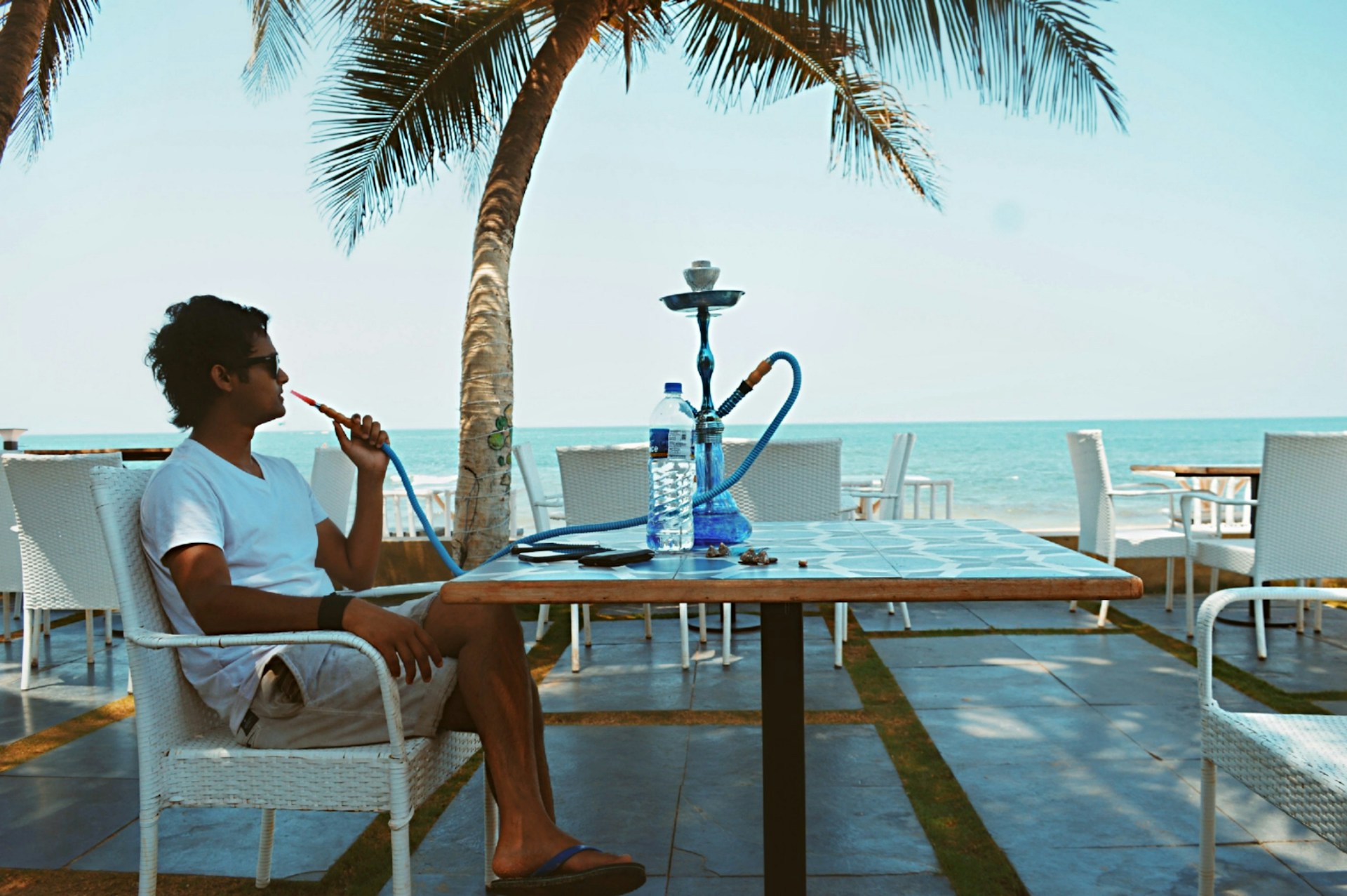 A person is sitting at a seaside cafe, enjoying a hookah. The setting includes palm trees, the ocean, and a blue sky. The individual, wearing a white shirt and shorts, relaxes in a wicker chair. A water bottle and a smartphone are placed on the table.