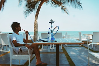 A person is sitting at a seaside cafe, enjoying a hookah. The setting includes palm trees, the ocean, and a blue sky. The individual, wearing a white shirt and shorts, relaxes in a wicker chair. A water bottle and a smartphone are placed on the table.