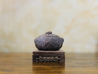 A rustic, textured pot sits atop an ornate wooden stand with intricate carvings. The backdrop is a soft, blurred light brown wall, complementing the earthy tones of the pot and the wooden surface.