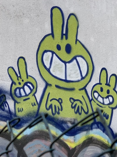 A graffiti mural featuring three stylized green cartoon rabbits with large, wide smiles and exaggerated facial features. The central rabbit is larger and positioned between two smaller rabbits. The artwork is outlined in blue and set against a white wall. The bottom of the image shows part of a black fence in the foreground, slightly blurred.