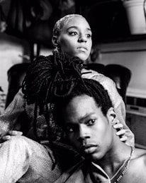 A black and white photograph of two individuals in an intimate pose. The person in the foreground has long dreadlocks and is looking off to the side with a contemplative expression. The person behind them, with a short hairstyle, gazes outward and embraces their shoulder. The setting appears casual, with soft lighting and blurred background elements.
