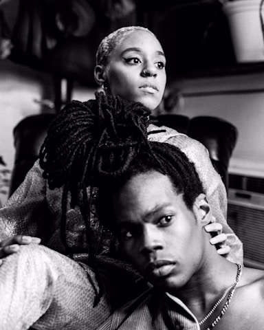 A black and white photograph of two individuals in an intimate pose. The person in the foreground has long dreadlocks and is looking off to the side with a contemplative expression. The person behind them, with a short hairstyle, gazes outward and embraces their shoulder. The setting appears casual, with soft lighting and blurred background elements.