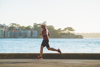 A person is jogging along a paved pathway near a body of water. In the background, there are several buildings and trees, with the horizon featuring a distant shoreline. The scene is well lit, indicating daylight, and the individual is dressed in athletic wear, including a cap and running shoes.