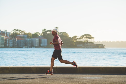 A person is jogging along a paved pathway near a body of water. In the background, there are several buildings and trees, with the horizon featuring a distant shoreline. The scene is well lit, indicating daylight, and the individual is dressed in athletic wear, including a cap and running shoes.