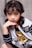A young girl with wavy dark hair poses with her chin resting on her hand. She is wearing a stylish white and black jacket with the text 'PARIS X90' and a black and gold shirt. Her expression is thoughtful, complemented by subtle makeup. The outfit includes striped sleeves and a floral-patterned skirt.