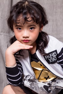 A young girl with wavy dark hair poses with her chin resting on her hand. She is wearing a stylish white and black jacket with the text 'PARIS X90' and a black and gold shirt. Her expression is thoughtful, complemented by subtle makeup. The outfit includes striped sleeves and a floral-patterned skirt.
