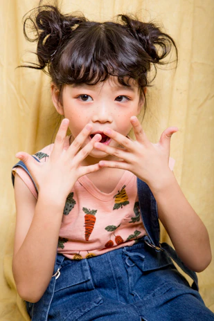 A young child with two messy buns in their hair is posing with their hands held up to their mouth in a playful manner. The child has no visible piercings or accessories and is wearing a light pink shirt with colorful carrot prints and denim overalls. The curtain-like yellow background contrasts with the outfit and hairstyle.