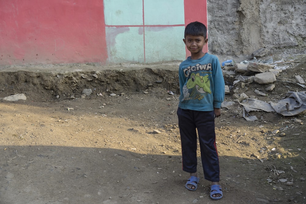 boy in blue sweater and black pants standing in front of teal and pink concrete wall