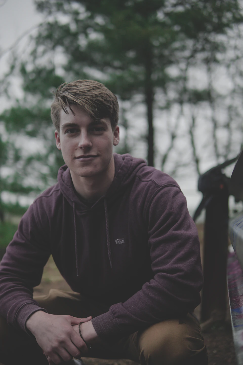 In the image, a young man is seated on a bench in what appears to be an outdoor setting. He is wearing a purple hoodie and brown pants, with his hands resting on his knees. The background features trees and a car parked nearby, suggesting that he might be in a park or a similar public space.

The image has a slightly blurred quality, giving it a dreamy, artistic feel. There are no discernible texts present in the photo.