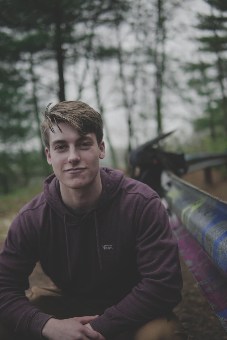 In a verdant forest, a young man sits on a wooden bench with his legs crossed and hands resting on his knees. He is wearing a purple hoodie and has short brown hair that falls neatly over his eyes. The background features tall trees with green leaves against an overcast sky.