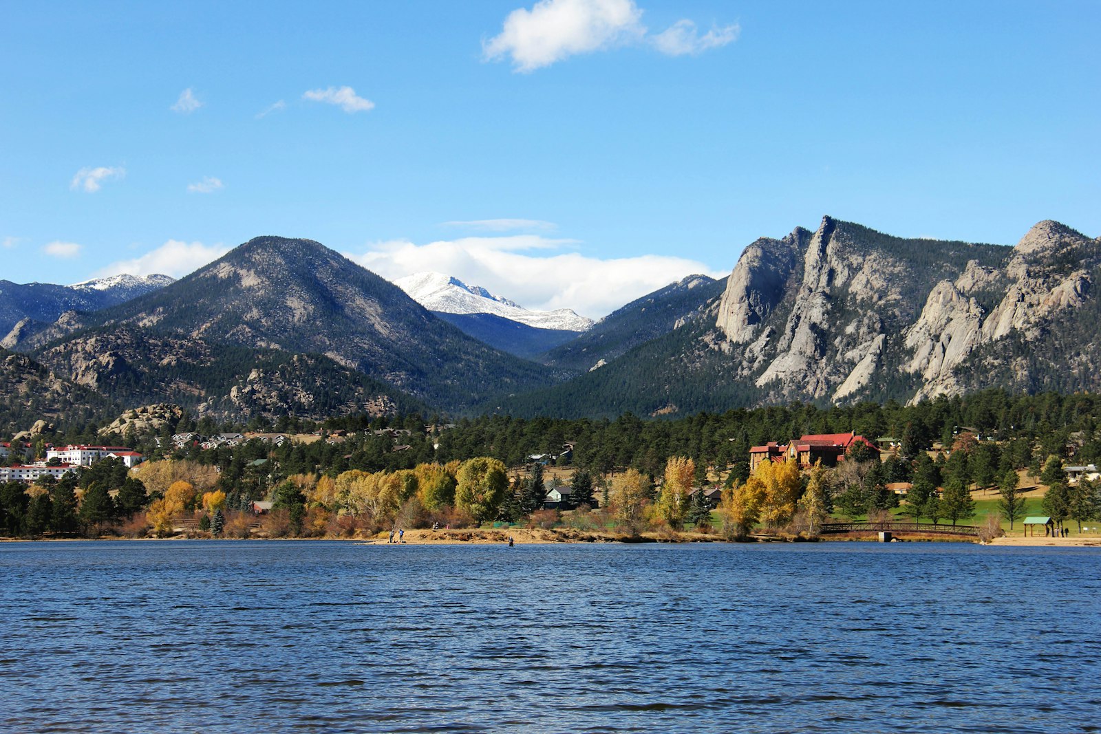 A wide shot of a lake in the foreground with a shoreline of autumn trees and buildings, backed by large rocky mountains including a distinctive flat-topped peak under a blue sky.