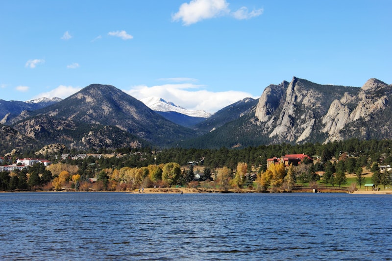 Blue water of Lake Estes in the foreground with golden autumn cottonwoods on the far shore, granite cliffs of Lumpy Ridge to the right and a snow-dusted Longs Peak in the distance