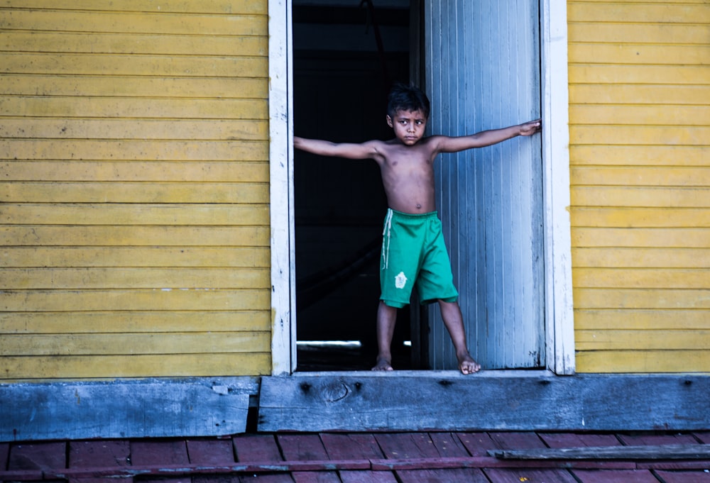 topless boy wearing green shorts standing beside door