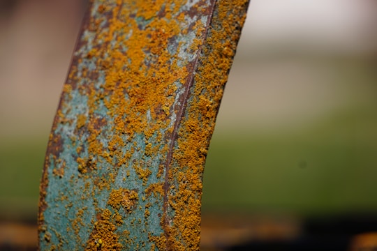 A close-up view of a rusted metal surface covered in a rich, orange-brown layer. The texture appears rough and uneven, with patches of blue-green paint visible underneath the rust. The background is blurred, consisting of neutral, earthy tones.