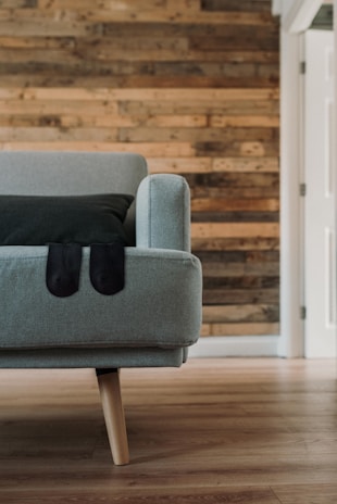 A modern, minimalist interior featuring a light gray couch with a dark cushion, set against a rustic wooden wall. The design exudes simplicity and elegance with a wooden floor complementing the wall.