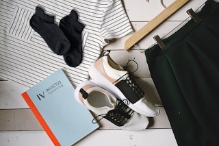 A flat lay arrangement featuring a striped shirt, a pair of black socks, a skirt on a hanger, a pair of stylish sneakers, and a book titled 'Invictus Tailoring' placed on a white wooden surface.